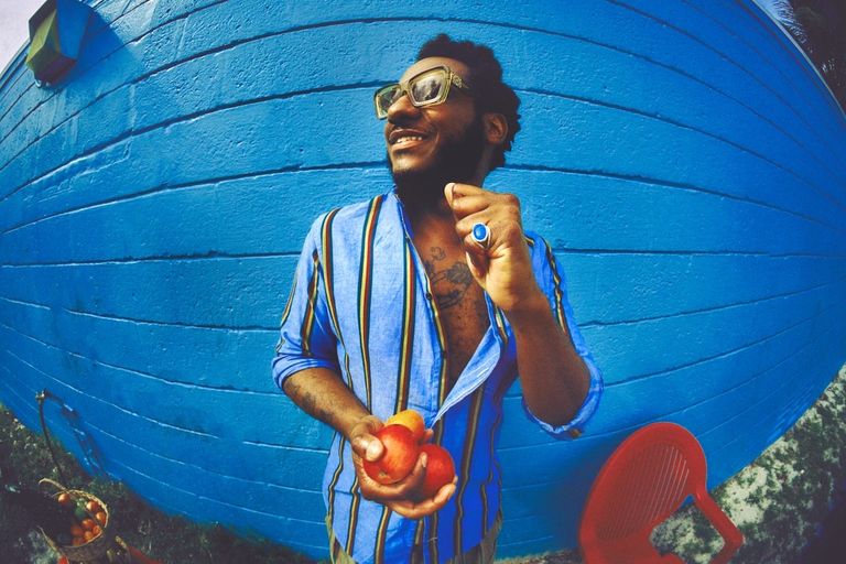 Soul singer Leon Bridges wearing sunglasses and a striped shirt, holding apples against a bright blue wall in a photo captured by Joshua Kissi.